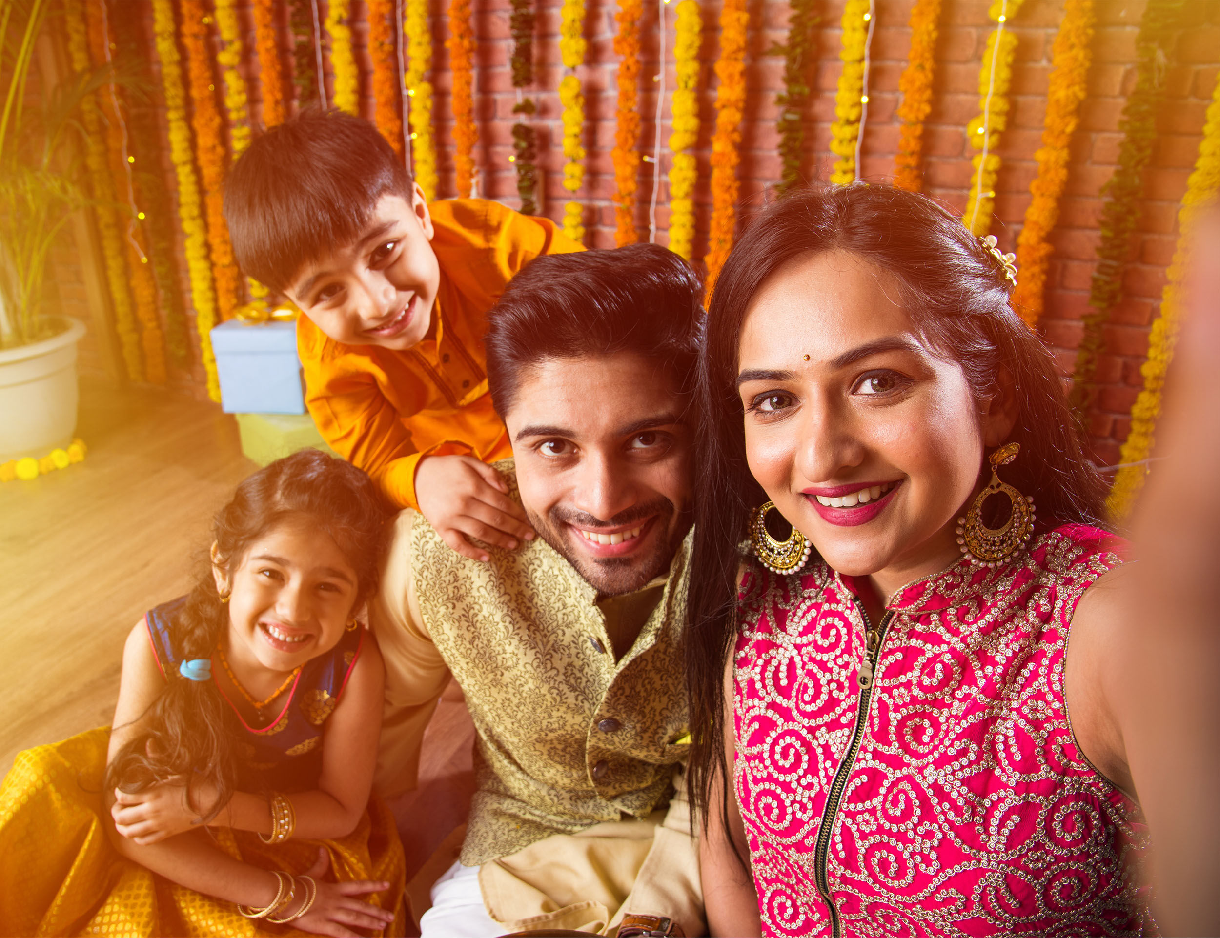 Happy Indian family celebrating Raksha Bandhan with kids, dressed in traditional festive attire and taking a joyful selfie in front of marigold flower decor
